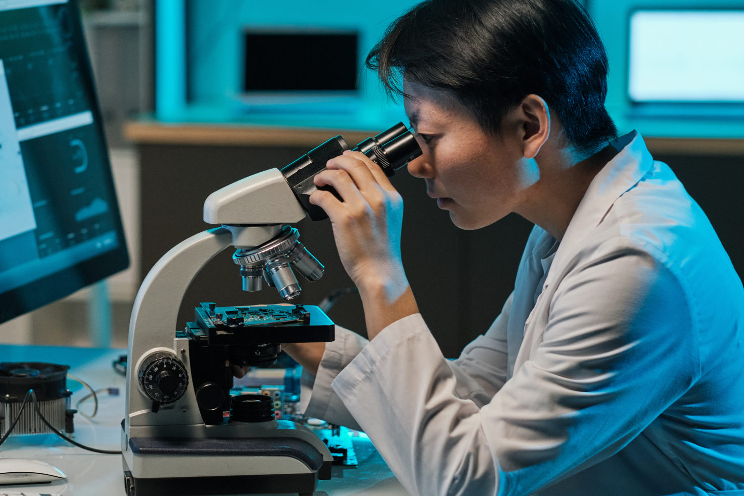 Asian female scientist looking in microscope during scientific investigation in front of two computer screens